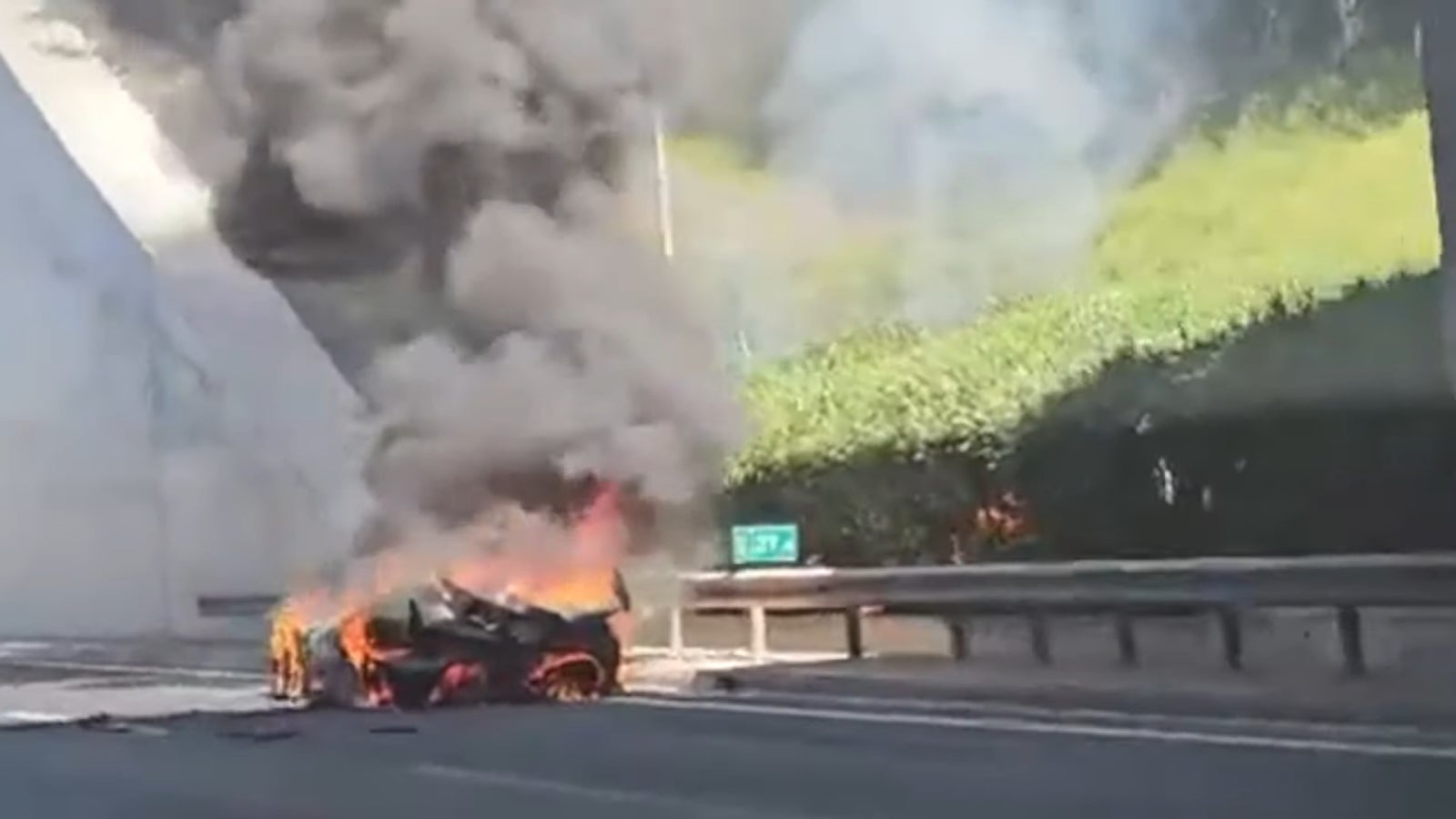 A screenshot showing a car on fire on the side of the road. 
